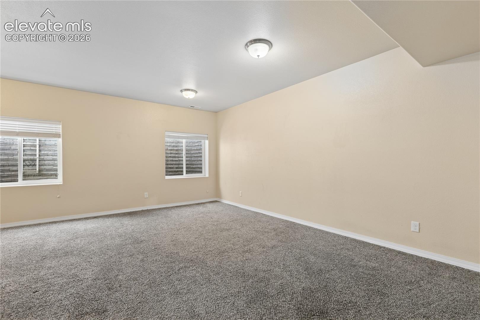 Spacious room featuring neutral wall paint, gray carpeting, dual windows with blinds, and flush-mount lighting fixtures