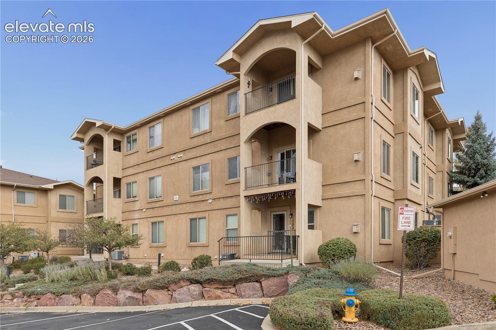 Multi-story stucco exterior featuring arched balconies with metal railings, varied roofline elevations, and native landscape beds with natural stone retaining walls