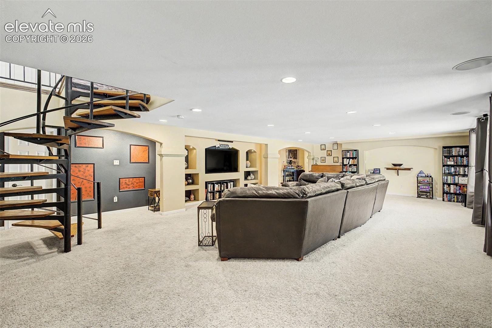 A dramatic spiral staircase connects this expansive lower-level living room to the floor above, where carpeted comfort, built-in bookshelves, and arched display niches create a true second living space for the home.