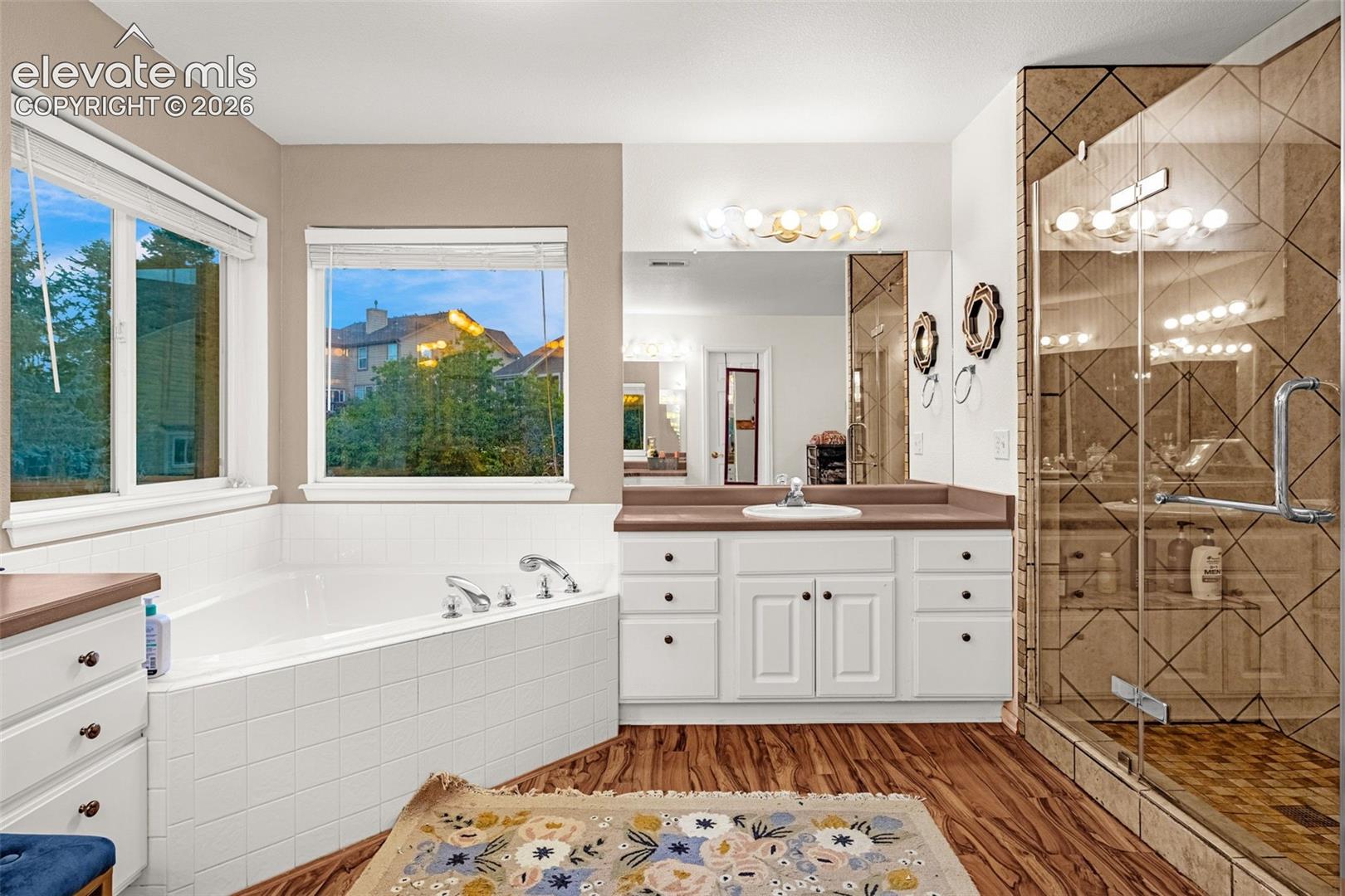 Wood-finish flooring with a white built-in tub, white vanity with a single basin, and a glass-enclosed shower with patterned wall tile