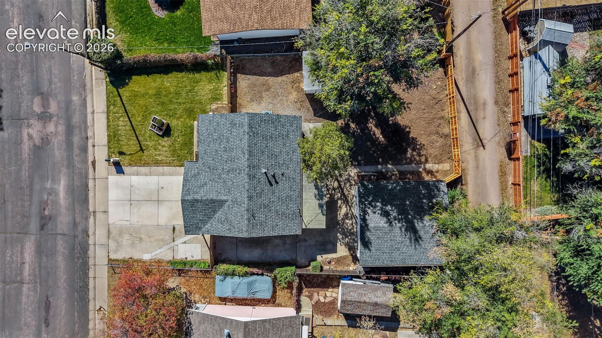 Aerial perspective featuring a shingled roof, concrete driveway, fenced perimeter, and mature trees on a level lot