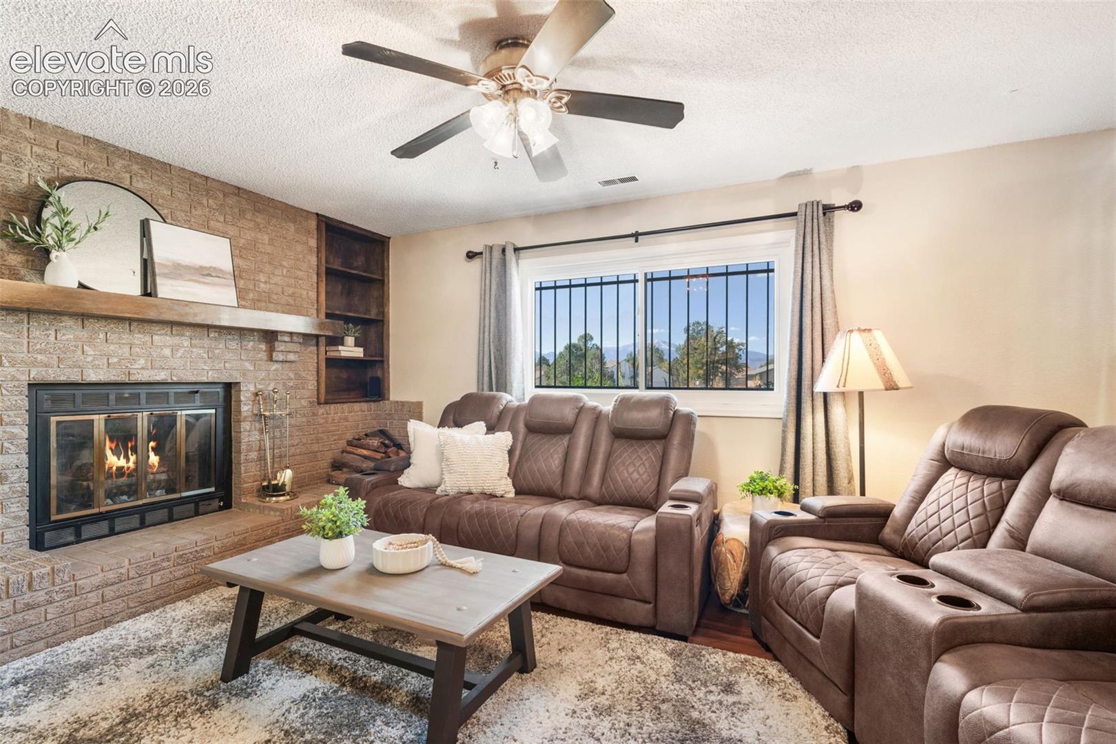 Floor-to-ceiling brick fireplace with a wood mantel, built-in shelving, ceiling fan with light fixture, and horizontal sliding window