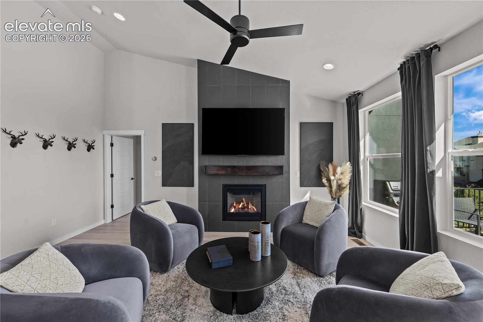 Vaulted ceilings featuring a black matte ceiling fan, floor-to-ceiling tile fireplace surround, wood-finish flooring, and large windows with recessed lighting