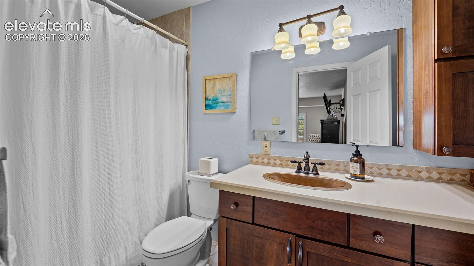 Bathroom featuring wood-finish cabinetry, an oval copper-tone vessel sink, neutral solid-surface countertop with decorative tile backsplash, and three-light wall-mounted vanity fixture