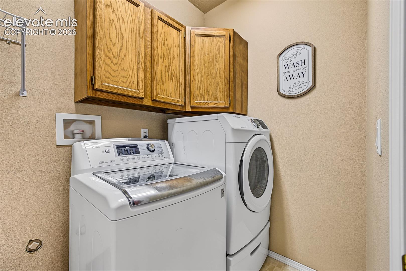 Private laundry room within the walkout basement bedroom suite, featuring a washer and dryer that stay with the home, along with overhead cabinetry for additional storage.