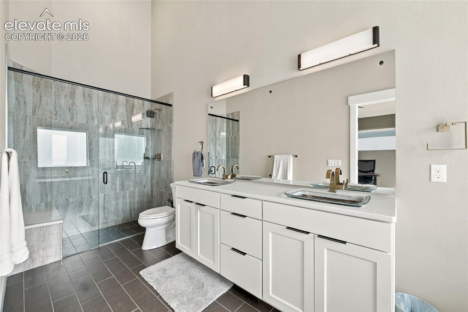 Dual-vanity cabinet with stone-finish countertop, under-mount sinks, gold-tone fixtures, large mirror, and glass-enclosed walk-in shower with tile surround