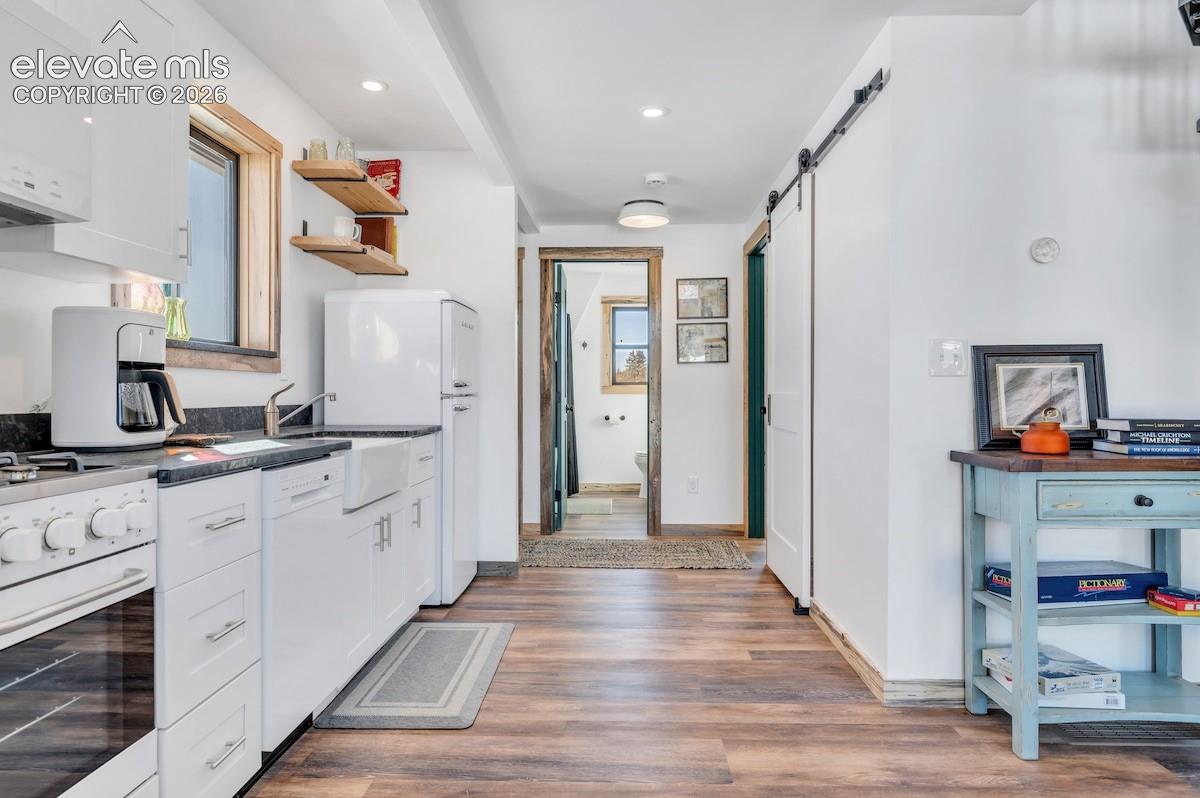 Kitchen featuring white shaker-style cabinetry, dark stone countertops, an apron-front sink, open wood shelving, and wood-finish flooring with a sliding barn-style door