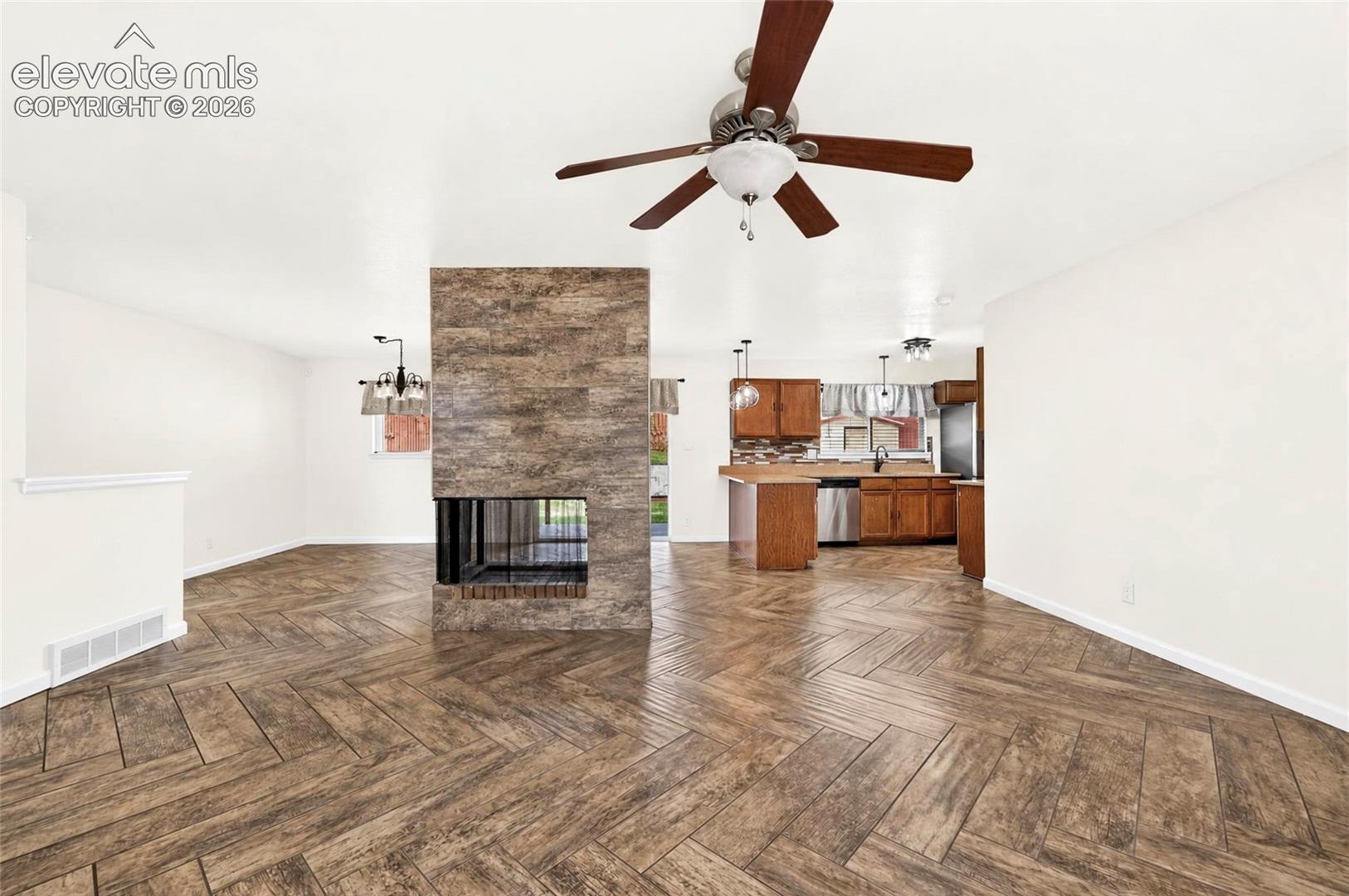 Herringbone-pattern wood-finish flooring, floor-to-ceiling stone-textured fireplace, wood cabinetry, stainless steel kitchen appliances, and ceiling fan