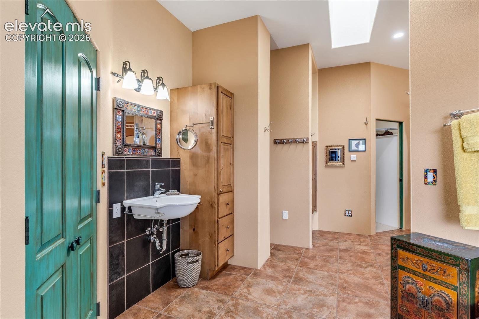Wall-mounted sink with chrome fixtures, floor-to-ceiling wooden storage cabinetry, black tile backsplash, decorative vanity mirror, and ceiling skylight