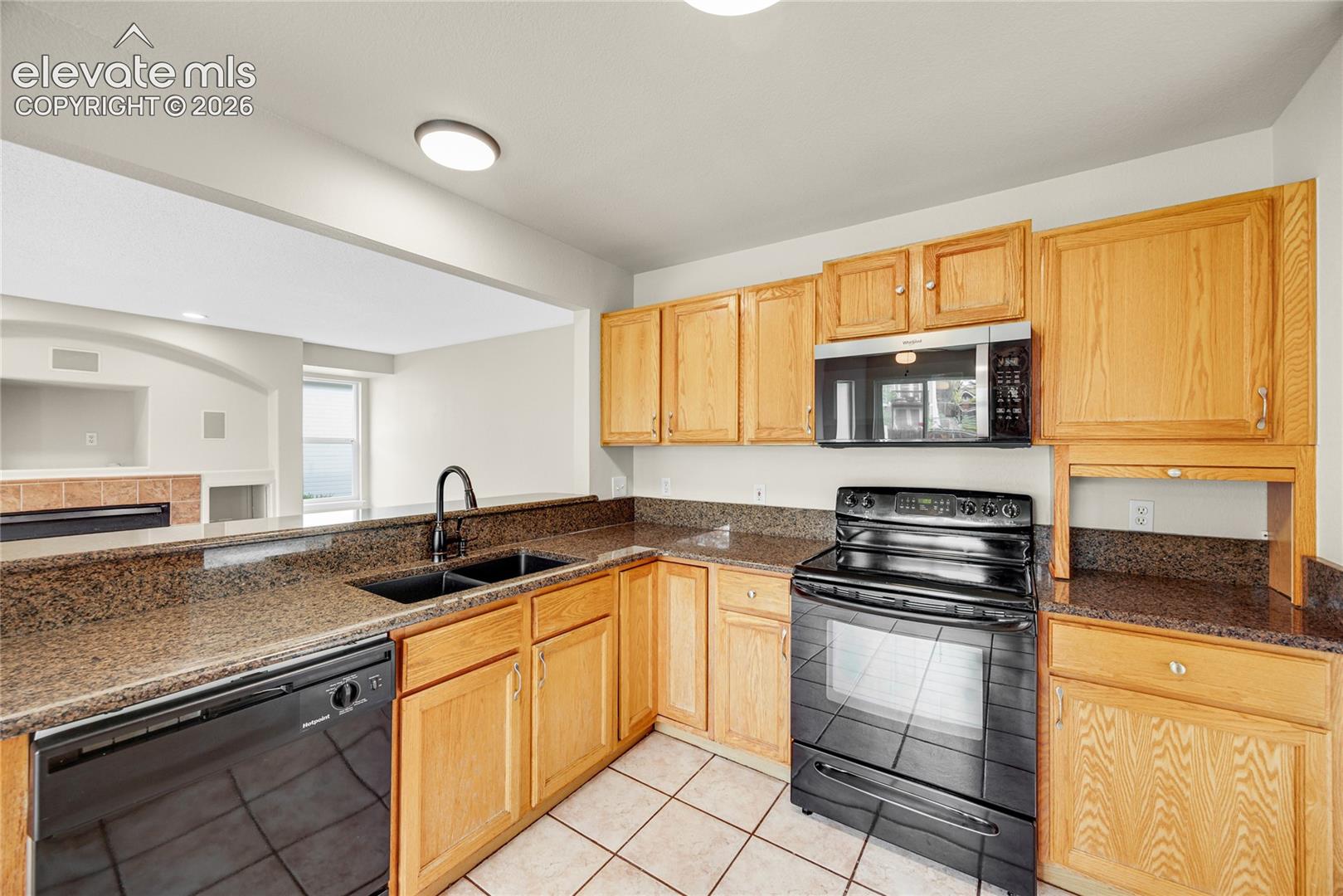 Kitchen featuring medium-tone cabinetry, stone-finish countertops, black built-in appliances, a dual-basin sink with a high-arch faucet, and ceramic tile flooring