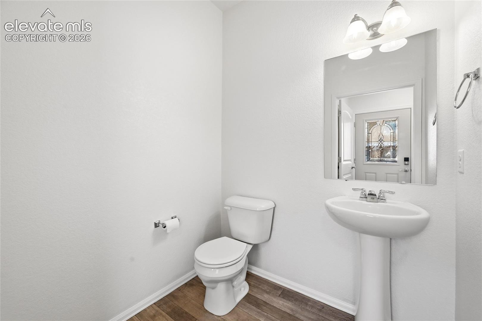 Powder room featuring wood-finish flooring, a white pedestal sink with chrome faucet, a rectangular vanity mirror, and a wall-mounted light fixture