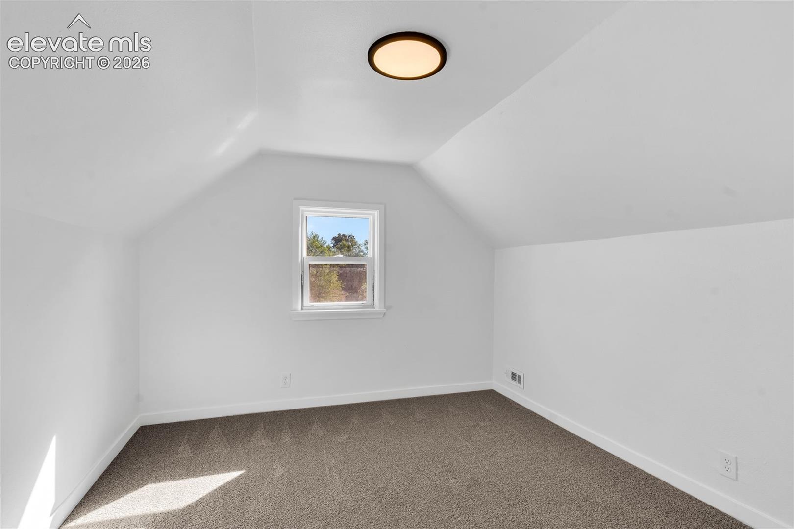 Interior space featuring slanted ceilings, neutral wall tones, carpeting, a single-hung window, and a flush-mount circular light fixture
