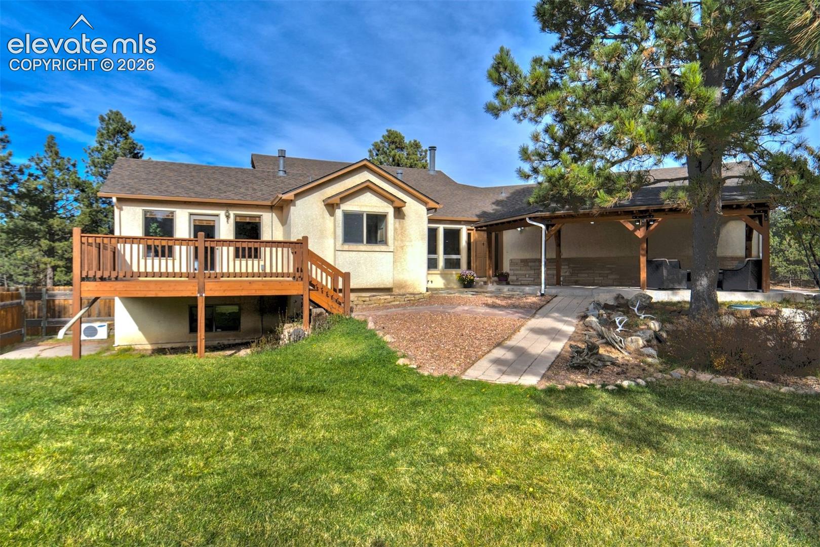 Exterior featuring a raised wood deck, a covered patio with stone-veneer accents, paved walkway, lawn, and mature trees