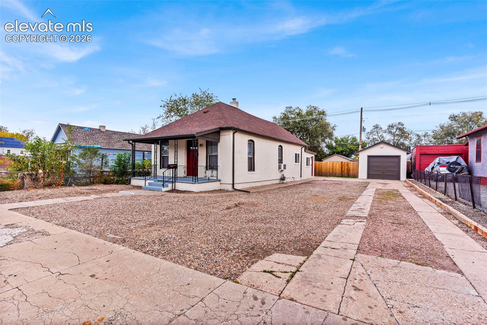 Single-story exterior featuring light-colored stucco siding, a hipped roof with asphalt shingles, an open front porch with metal railings, and a detached single-car garage