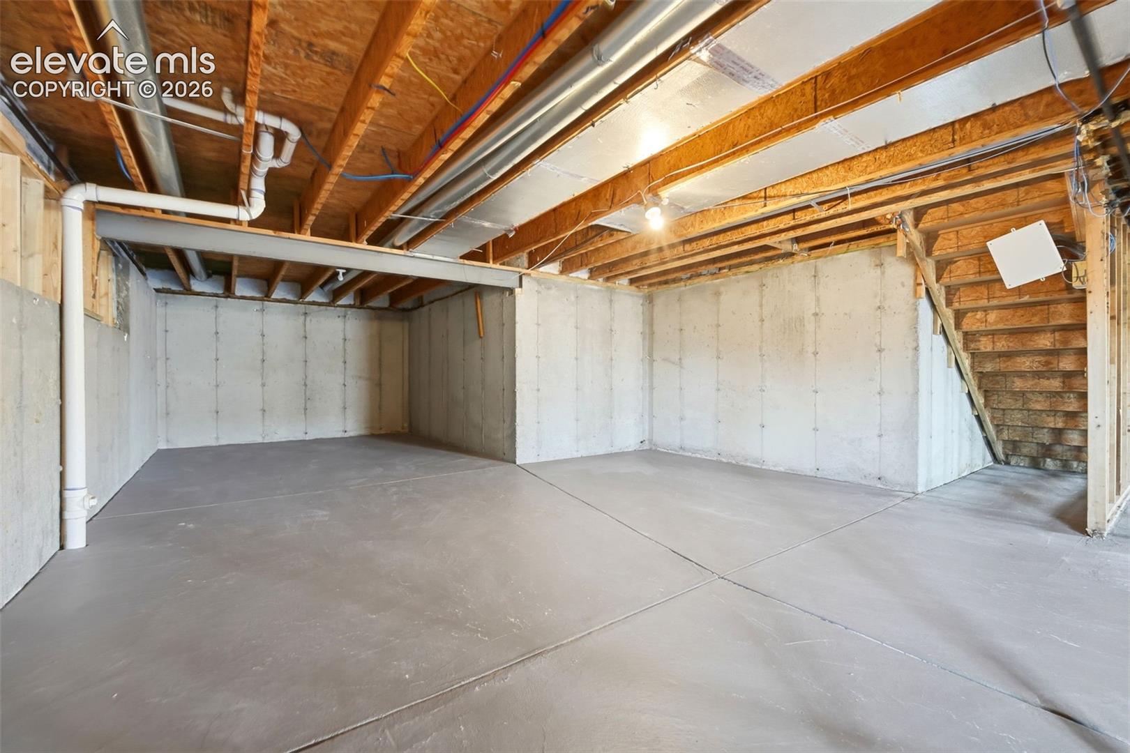 Unfinished basement with concrete flooring and exposed ceiling joists, featuring a visible wooden staircase