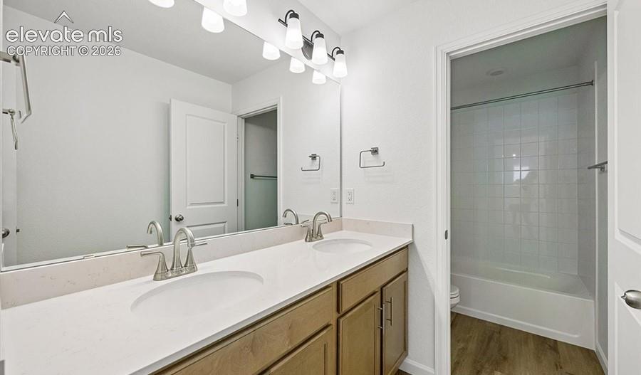 Dual-sink vanity with light-tone wood cabinetry, solid surface countertop, polished chrome fixtures, full-width mirror, and a tub-shower combination featuring white tile surround