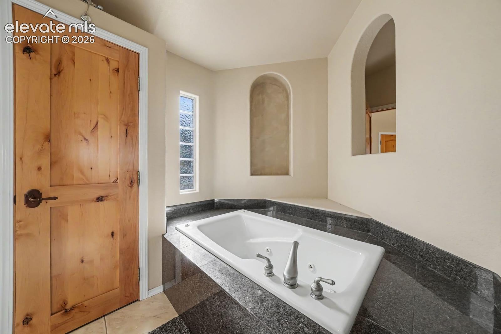 Soaking tub with integrated jets and stone-surround deck, featuring an arched recessed wall niche, a second arched cutout, and a natural wood-finish door with oil-rubbed bronze hardware