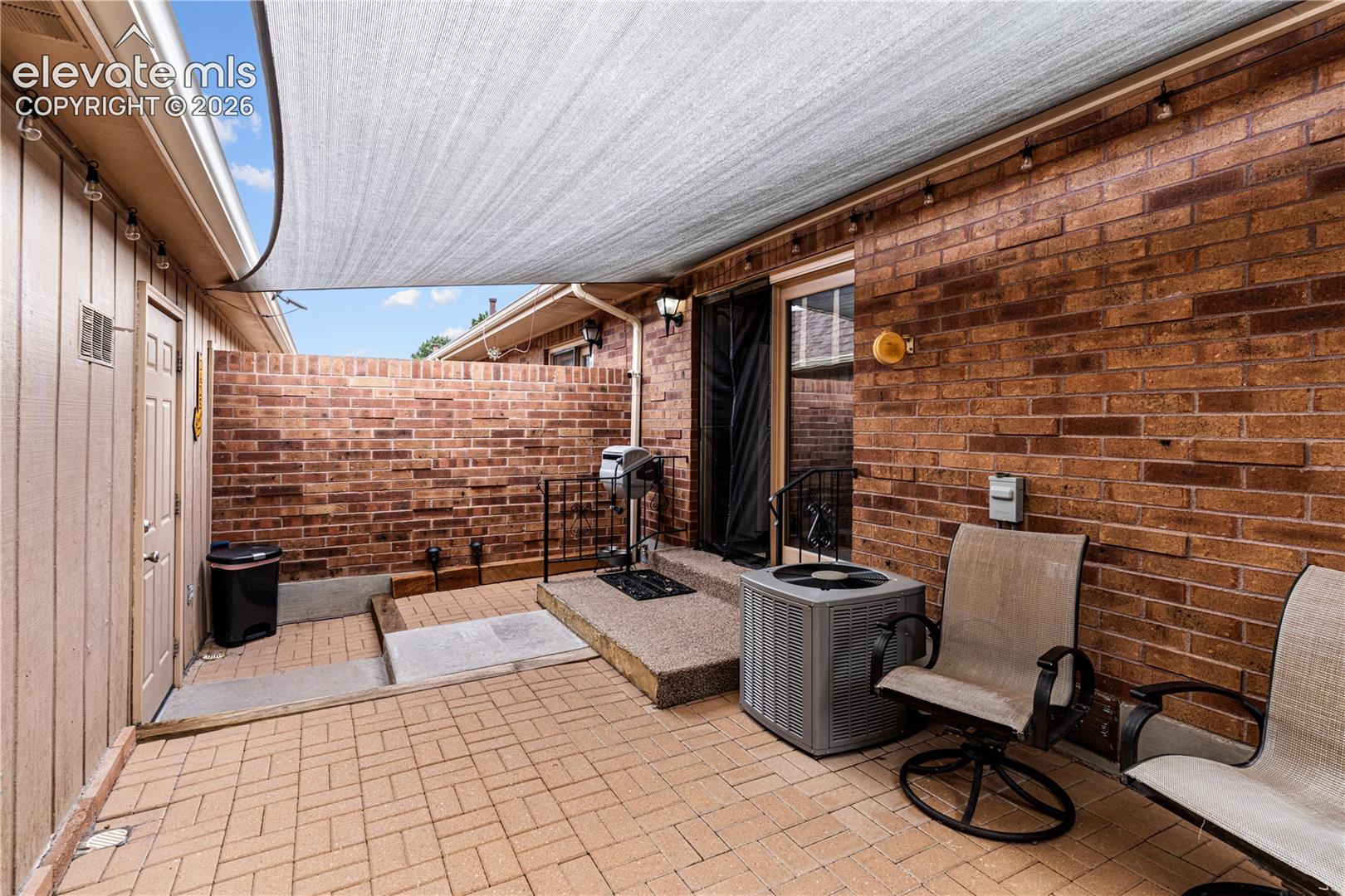 Enclosed patio featuring brick masonry walls, paver stone flooring, a multi-tiered concrete stoop, and an overhead shade sail