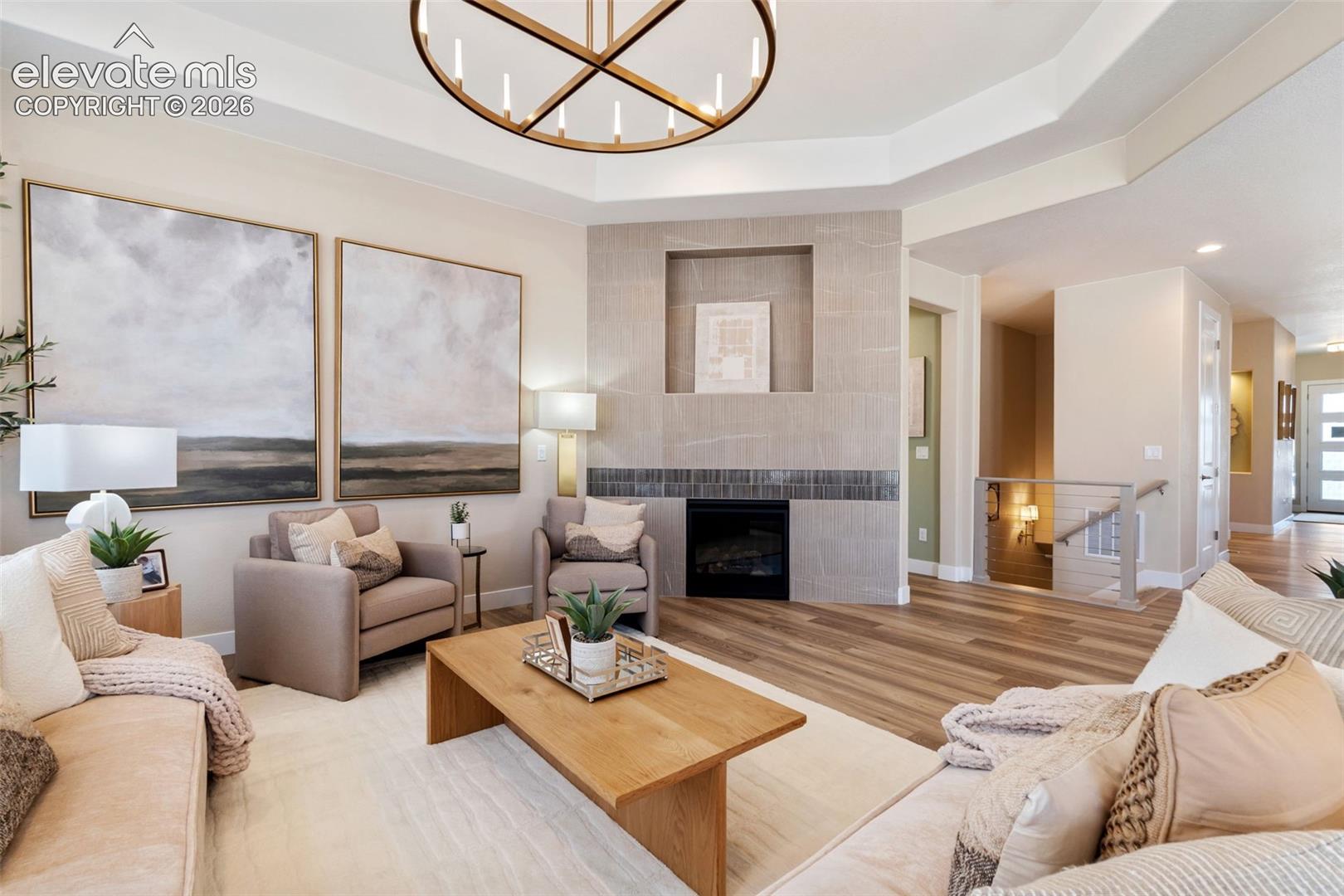 Tray ceiling featuring a large circular chandelier, fireplace with neutral stone-style surround, built-in display niche, and wood-finish flooring
