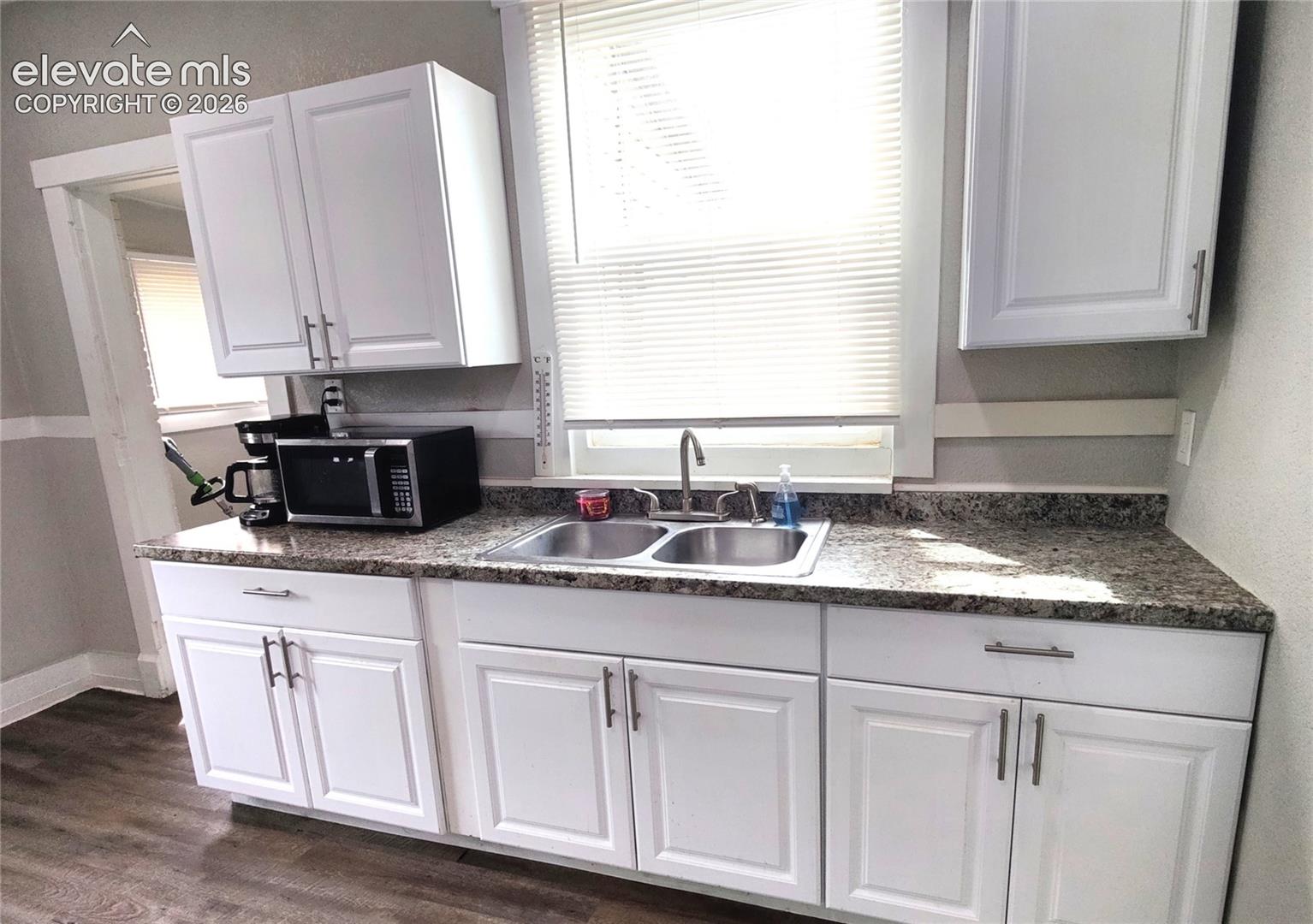 White shaker-style cabinetry, double basin stainless steel sink, speckled countertop, and wood-finish flooring