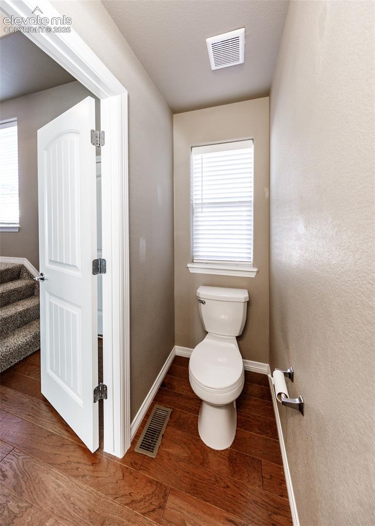 Wood-finish flooring, white multi-panel interior door with brushed nickel hardware, and window with white horizontal blinds