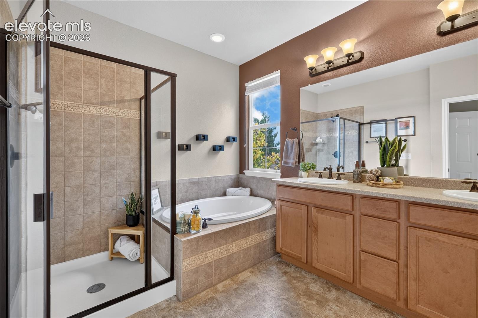 Bathroom featuring a dual-sink vanity with wood cabinetry, a tiled-surround soaking tub, a glass-enclosed shower with tiled walls, and a large wall-mounted mirror, Window for lots of light!