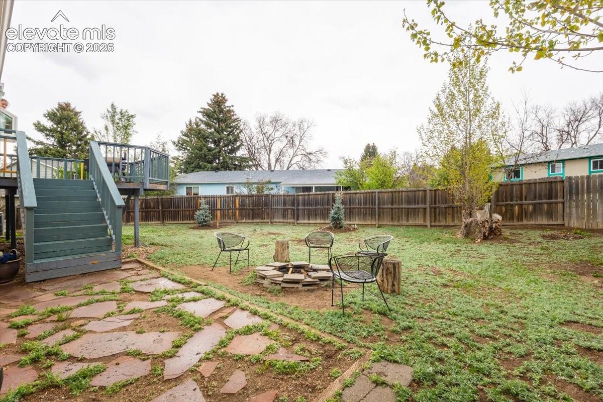 Spacious backyard featuring a wooden privacy fence, a multi-level wooden deck with stairs, a flagstone patio, a fire pit, and scattered trees