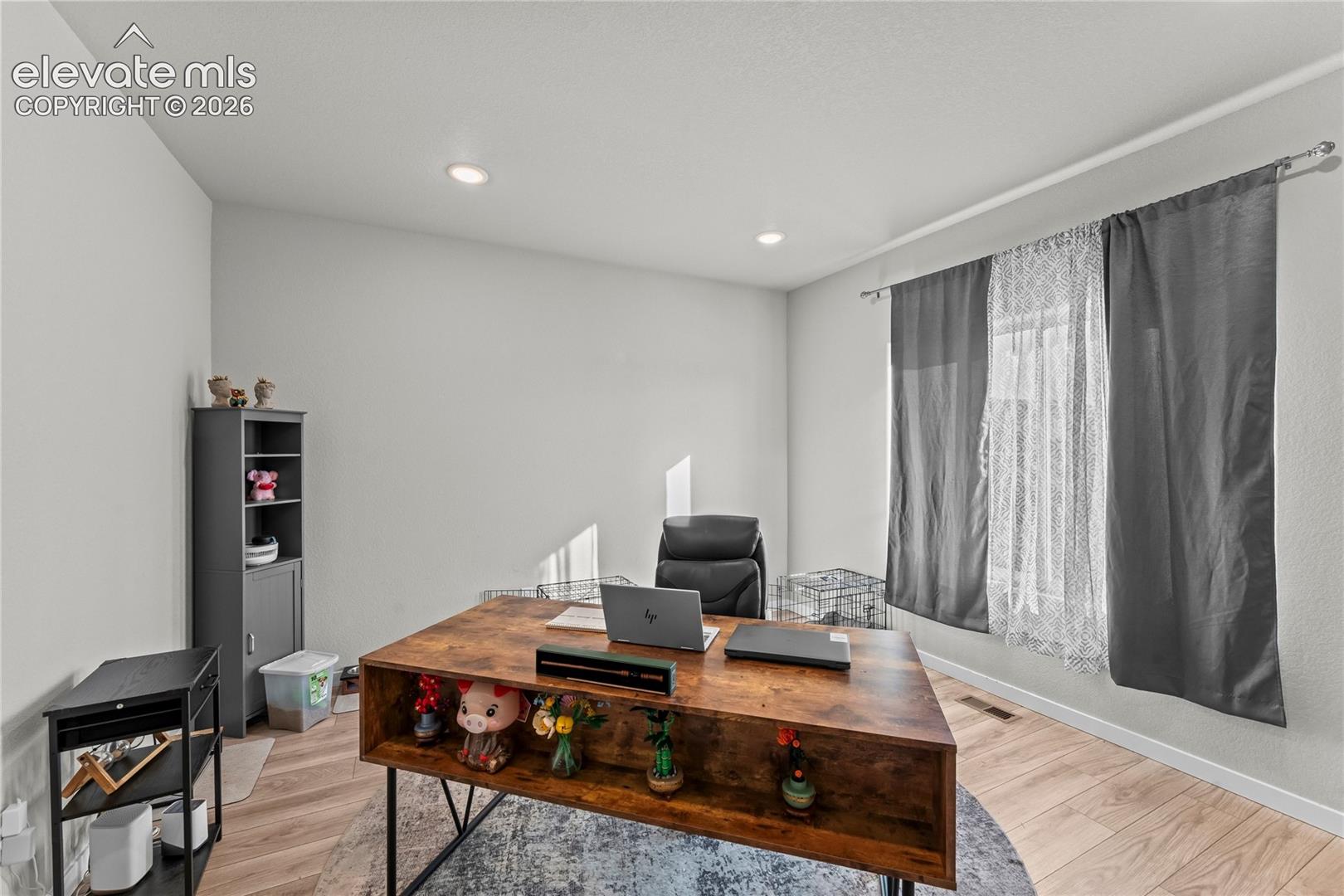 Dedicated home office featuring warm wood-finish flooring, a spacious work desk with integrated storage shelving, recessed ceiling lighting, and large windows dressed with layered gray and sheer curtains that provide natural illumination and privacy control