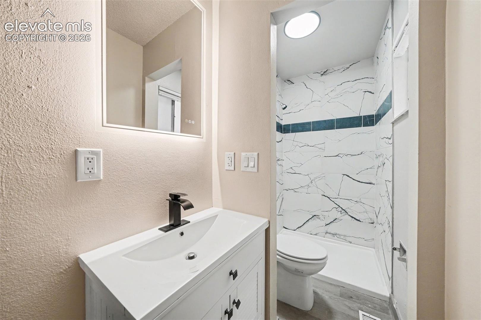 Bathroom featuring a white vanity with a rectangular sink, matte black faucet, wall-mounted backlit mirror, and a walk-in shower with stone-patterned tile surround
