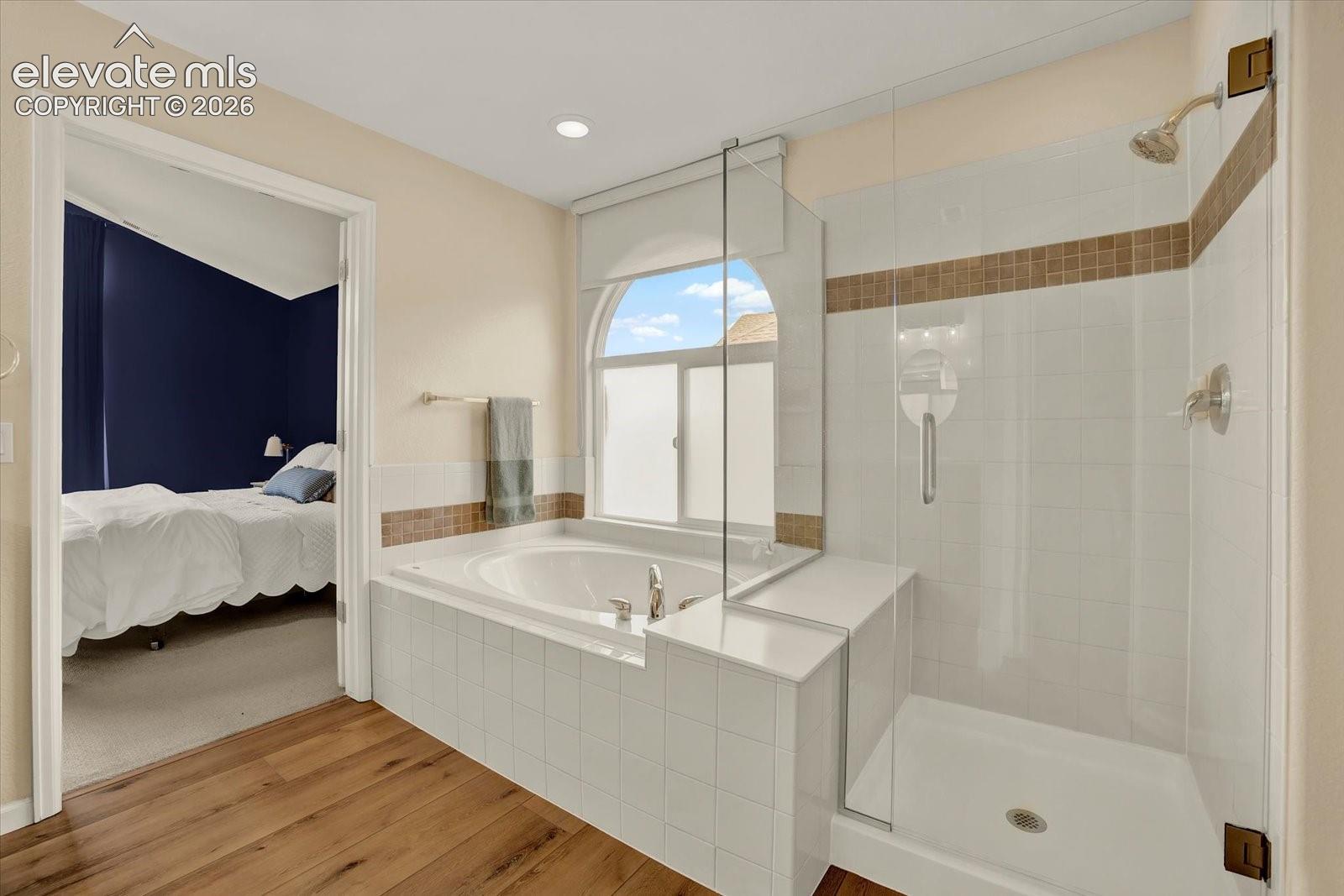 Soaking tub with a tile surround and arched window, glass-enclosed walk-in shower, and wood-finish flooring