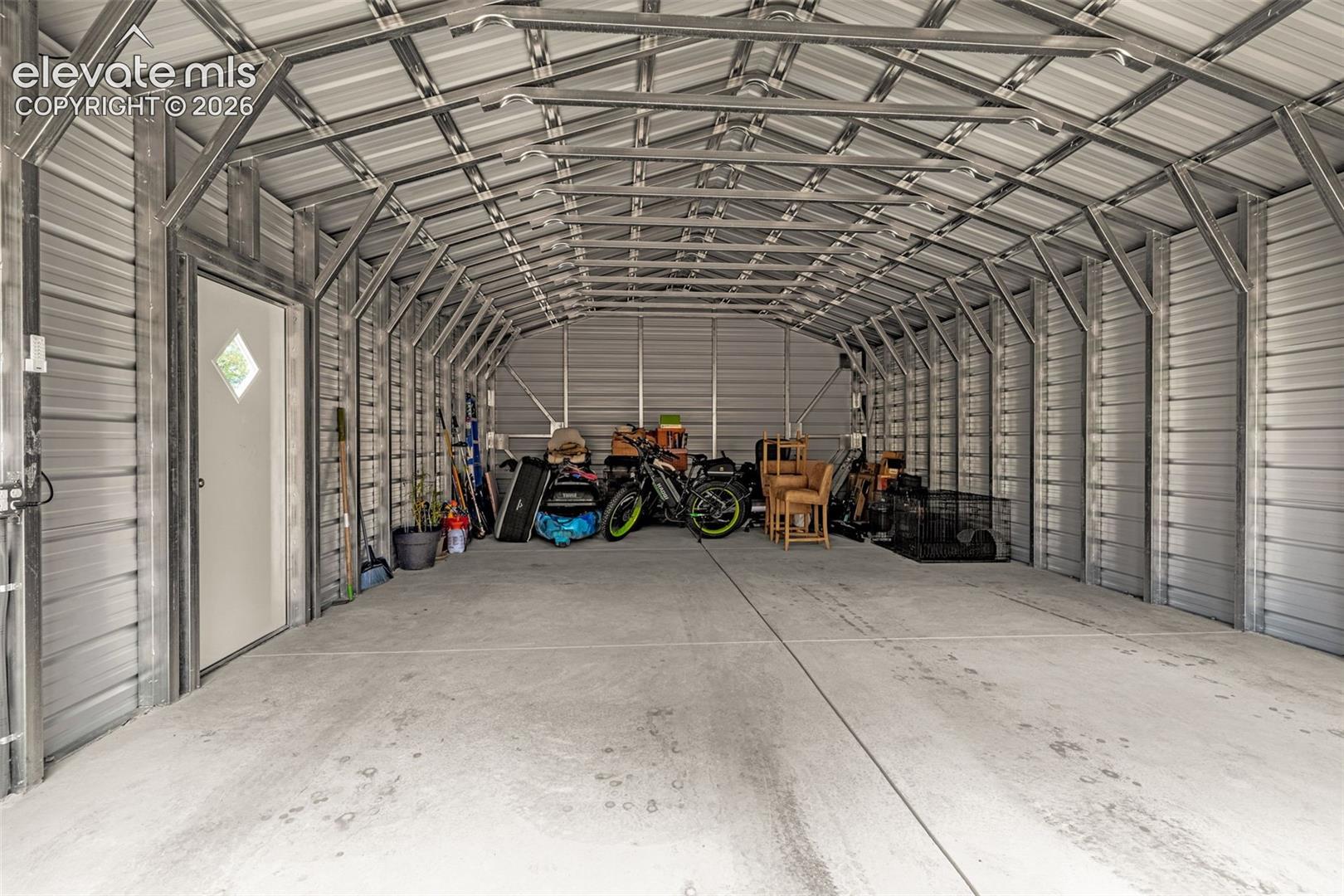 Enclosed metal structure featuring a concrete foundation, exposed steel truss framing, corrugated metal walls and roof, and a single pedestrian access door