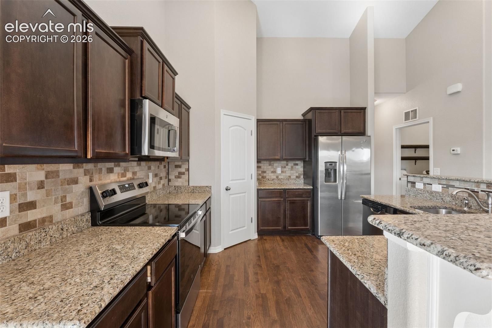 Kitchen featuring dark wood cabinetry, stainless steel appliances, a multi-tone tile backsplash, and light speckled countertops