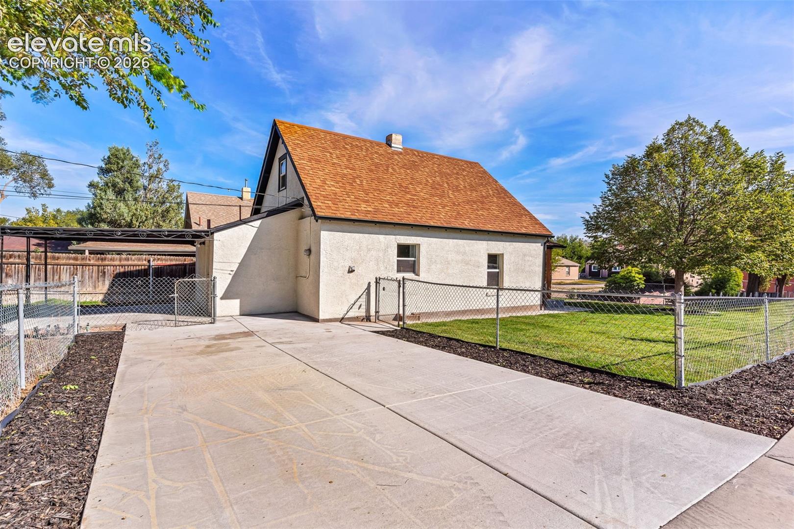 Stucco exterior featuring a pitched shingle roof, concrete driveway, covered parking structure, and chain-link fenced yard