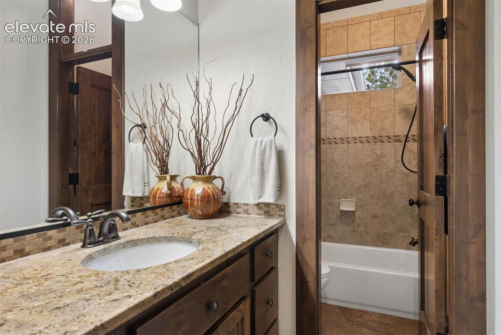 Bathroom vanity featuring a stone-composite countertop with an integrated undermount sink, custom wood cabinetry, dark-finish hardware, and tiled tub surround