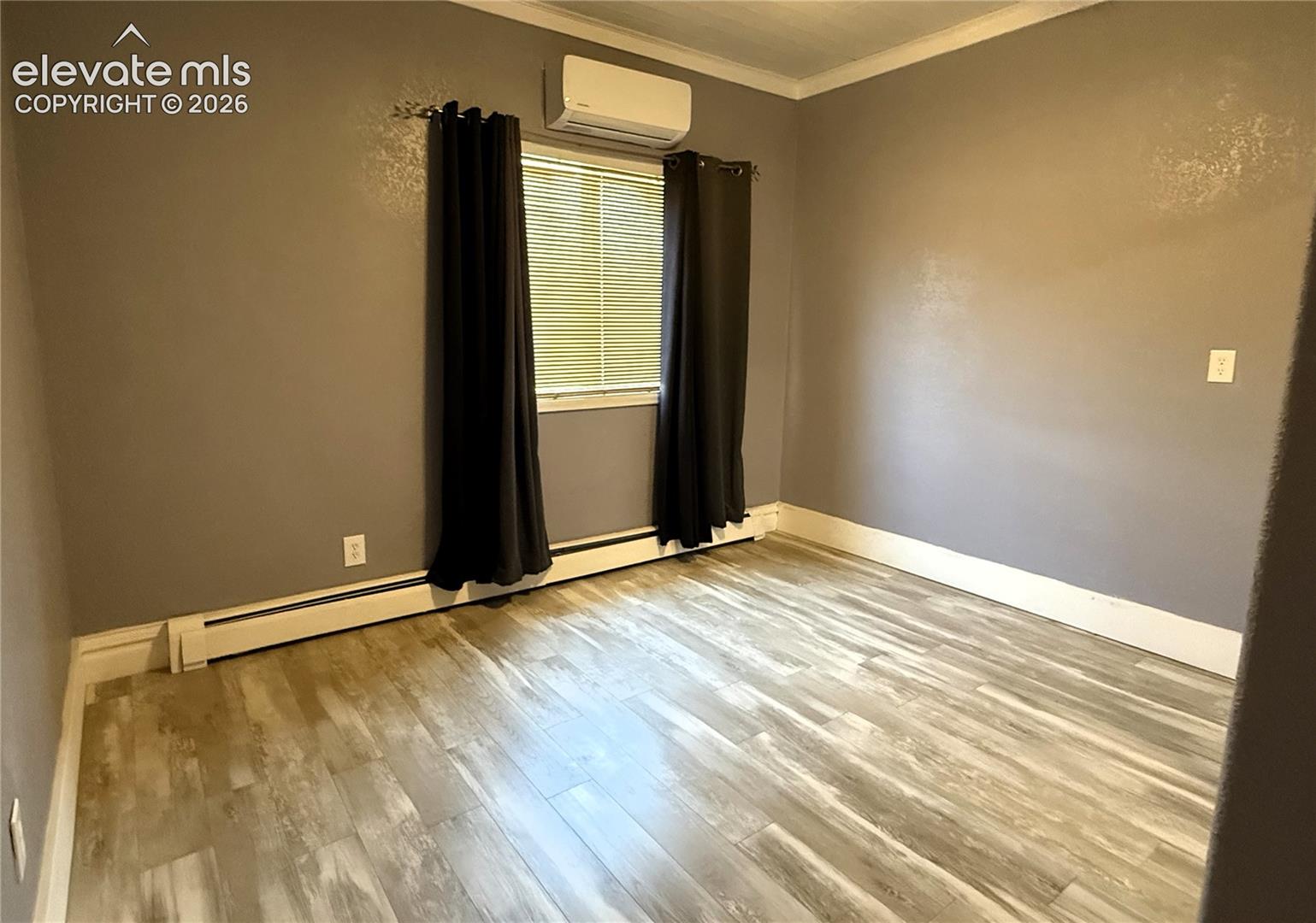 Neutral-toned room featuring wood-finish flooring, a wall-mounted mini-split HVAC unit, and a window with horizontal blinds