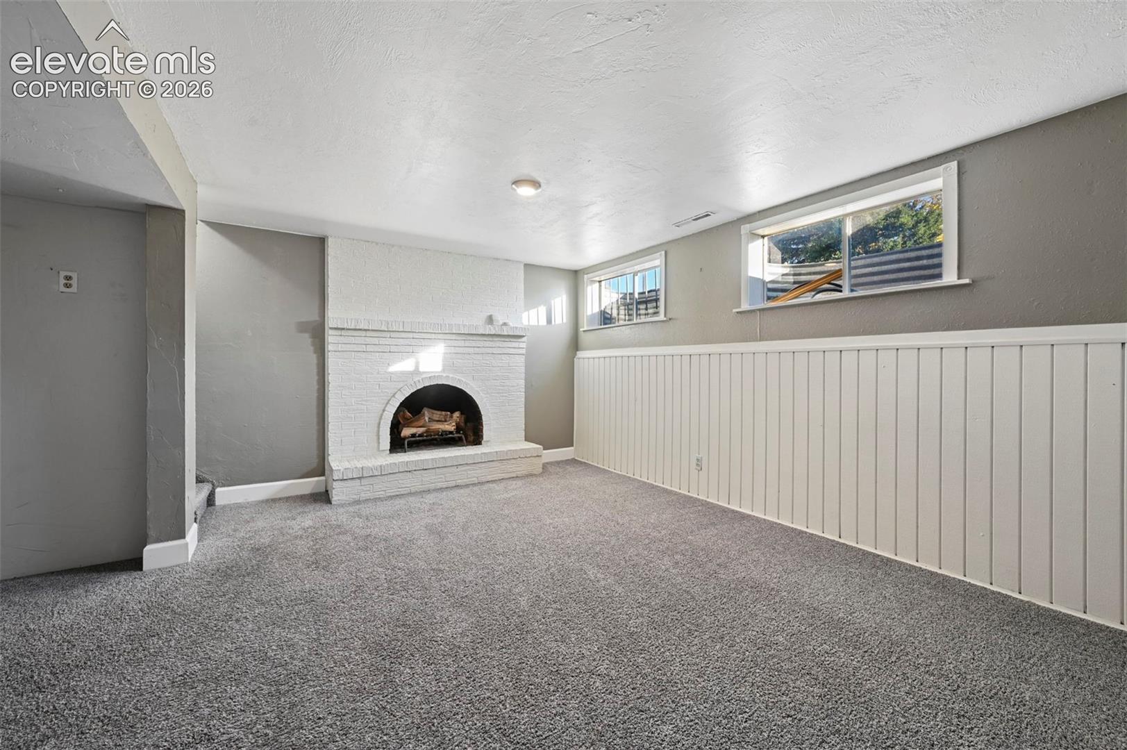 Unstaged family room in Basement living room with cozy painted brick fireplace with an arched firebox, white vertical wall paneling, and wall-to-wall carpeting!