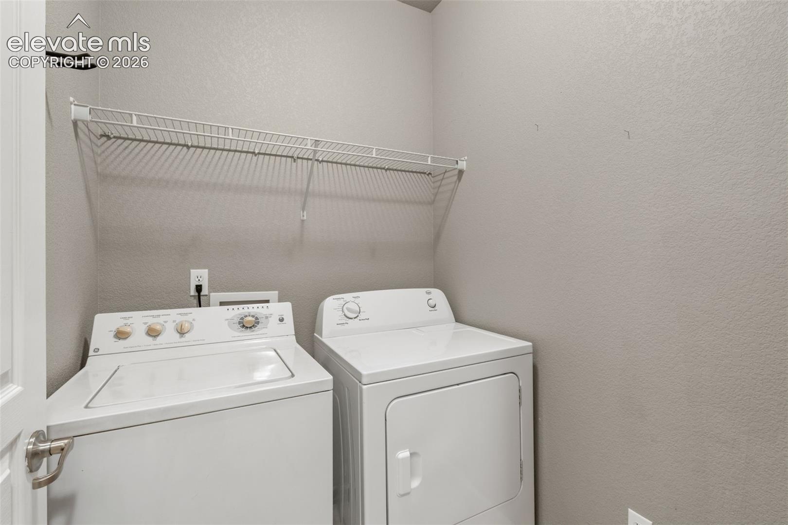 Laundry area featuring white top-load washer and dryer, mounted wire shelving, and neutral wall finish