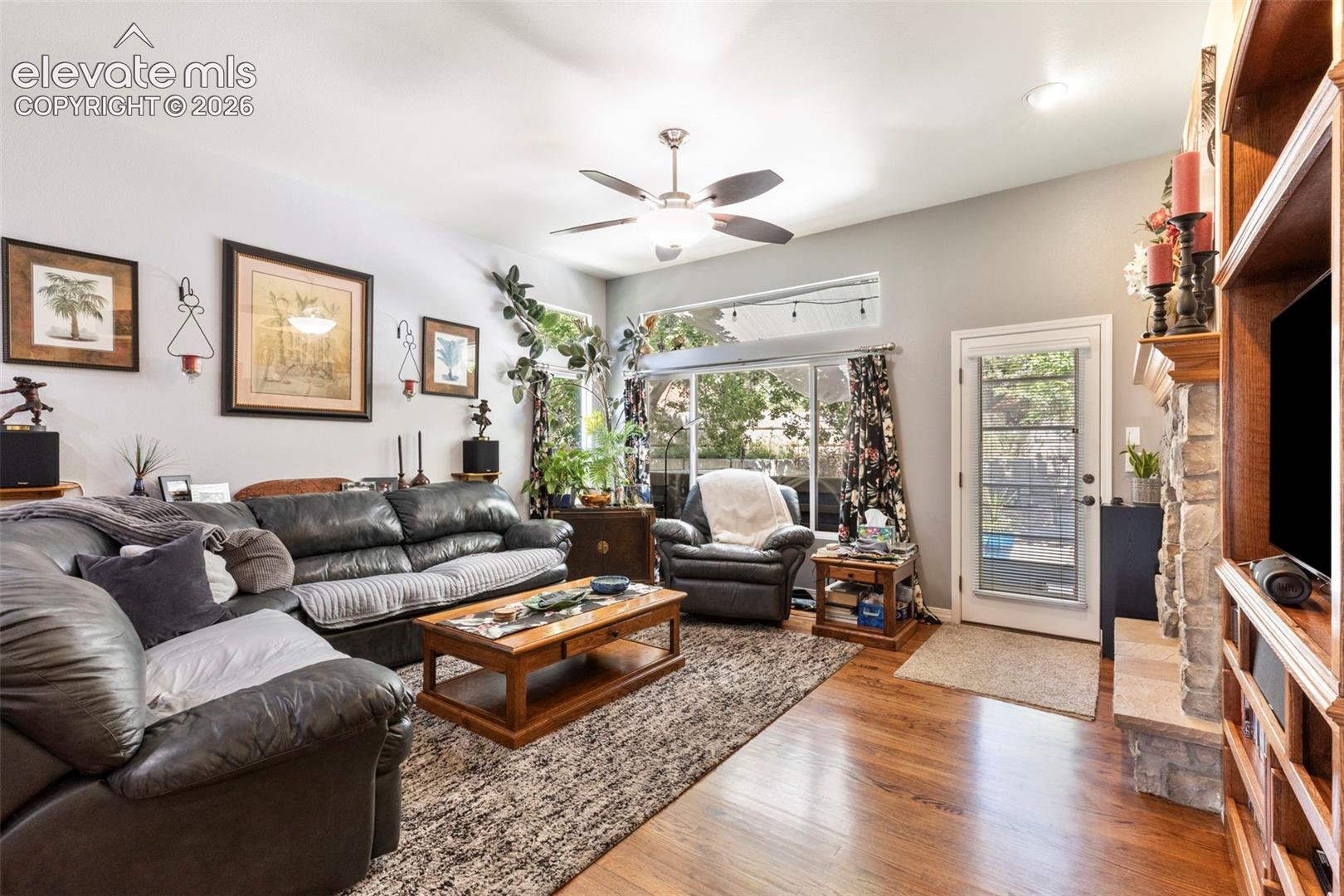 Hardwood flooring throughout, a ceiling fan fixture, a built-in entertainment center, and a stone-faced fireplace with a wood mantel