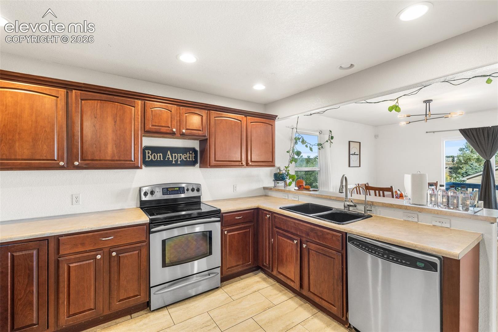 Kitchen featuring wood cabinetry, stainless steel appliances, neutral solid-surface countertops, dual-basin undermount sink, and tile flooring