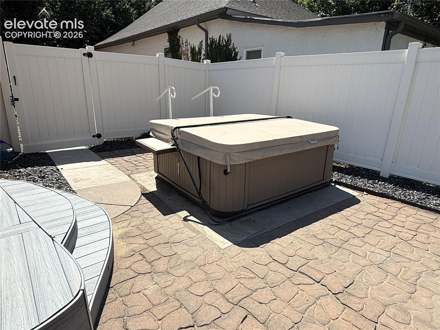 Outdoor spa area featuring a textured concrete paver patio, composite deck steps, and a white vinyl privacy fence