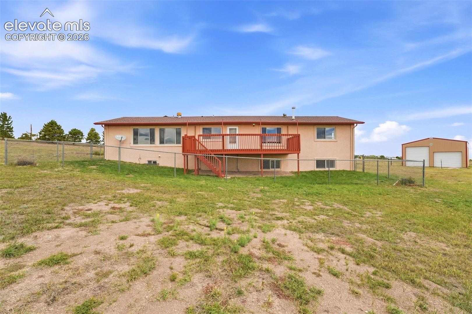 Ranch-style exterior with a stucco finish, a raised wooden deck, a lower-level concrete patio, a chain-link fence, and a detached two-door outbuilding