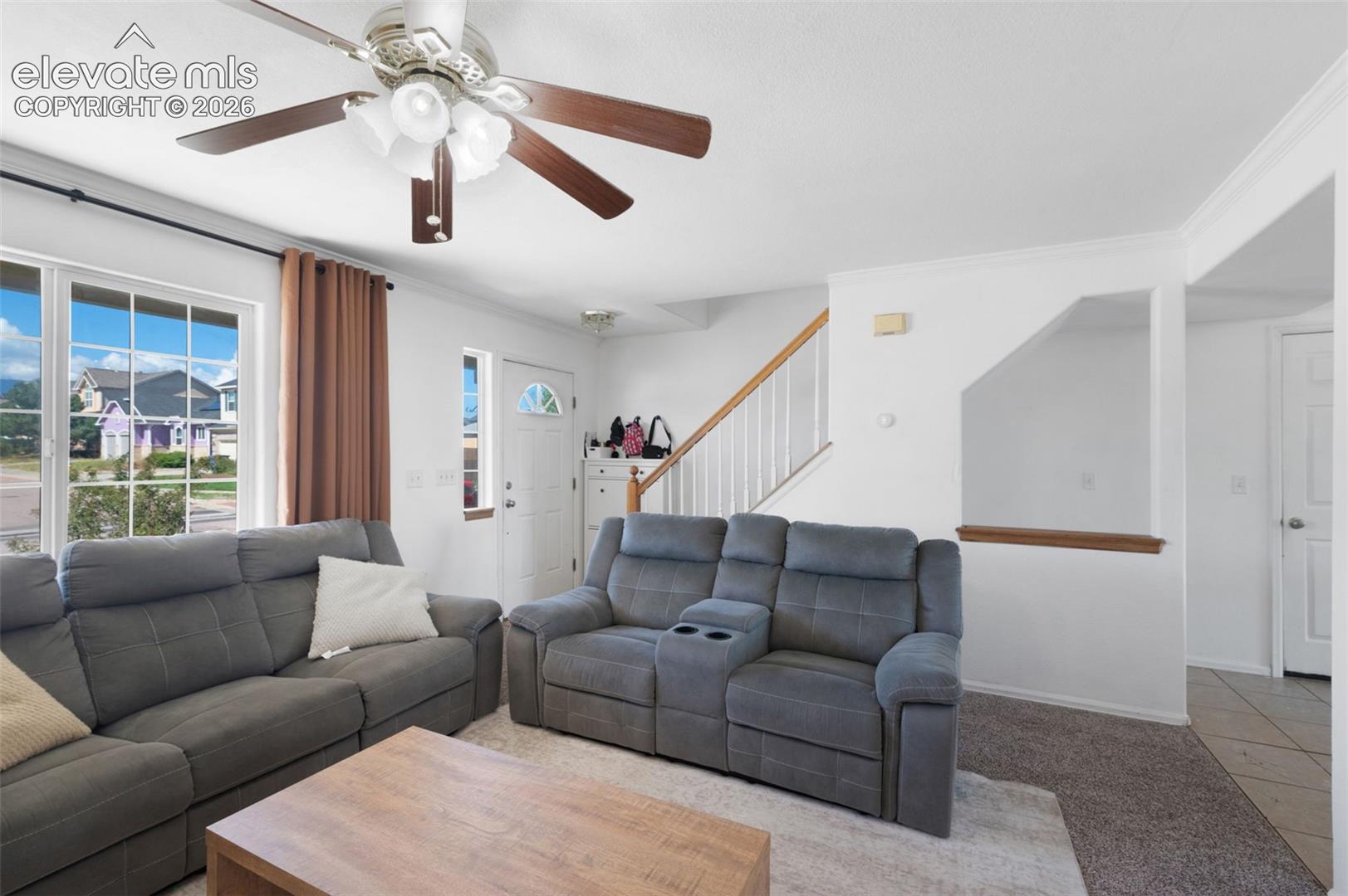 Living area featuring a ceiling fan with integrated lighting, large window with grid pattern, and neutral carpet flooring