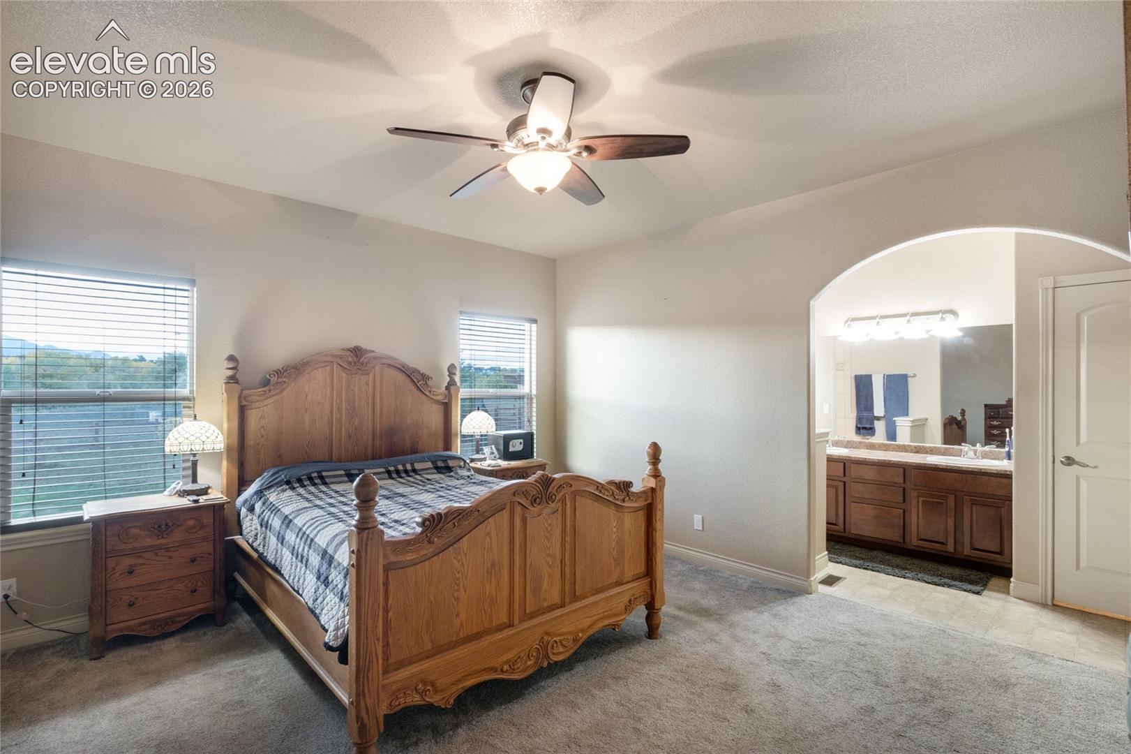 Room with neutral wall color, plush carpeting, ceiling fan with integrated lighting, arched entryway, and natural light via multiple windows; attached bathroom in the background, double vanity