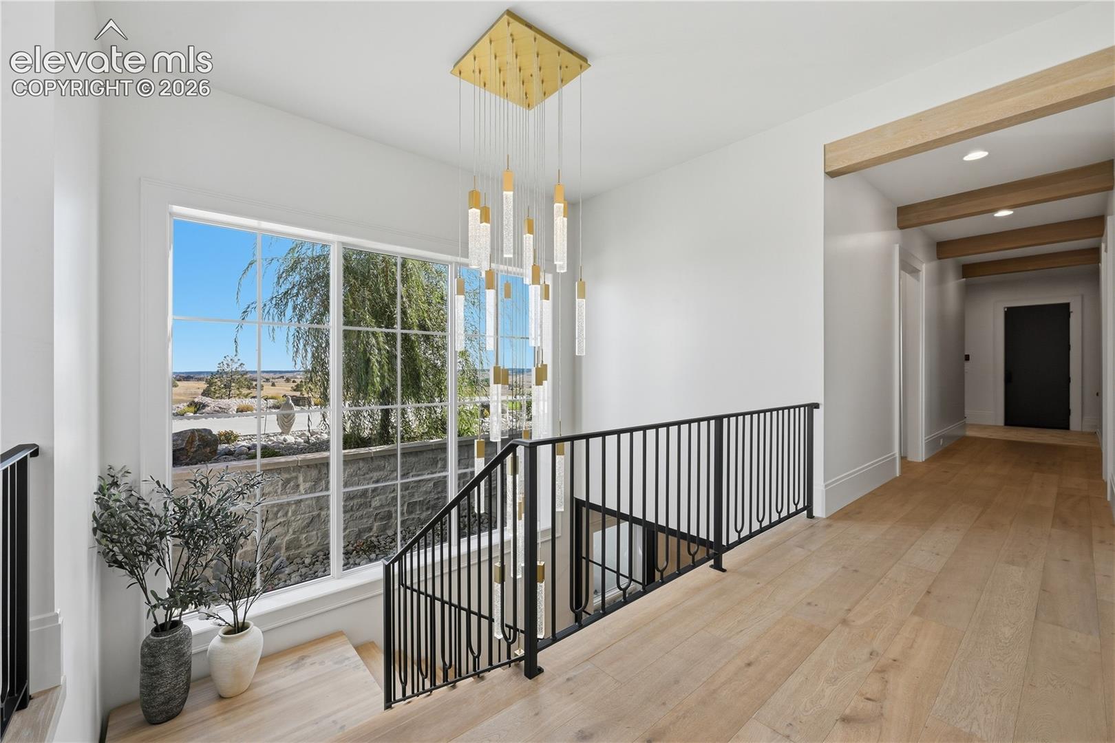 Multi-level foyer featuring wood-finish flooring, a tiered gold-finish chandelier with cylindrical glass pendants, black metal railing, and exposed ceiling beams