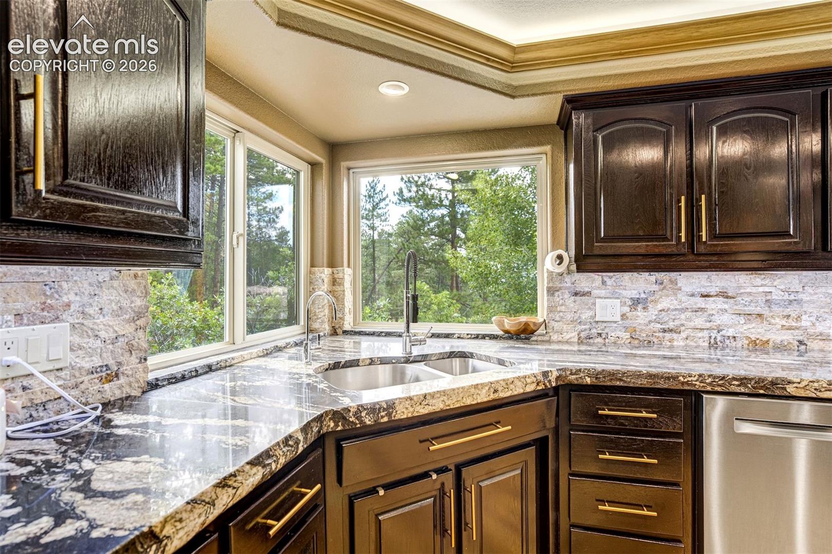 Dark wood cabinetry with gold-tone hardware, granite countertops, tile backsplash, and a stainless steel double basin sink