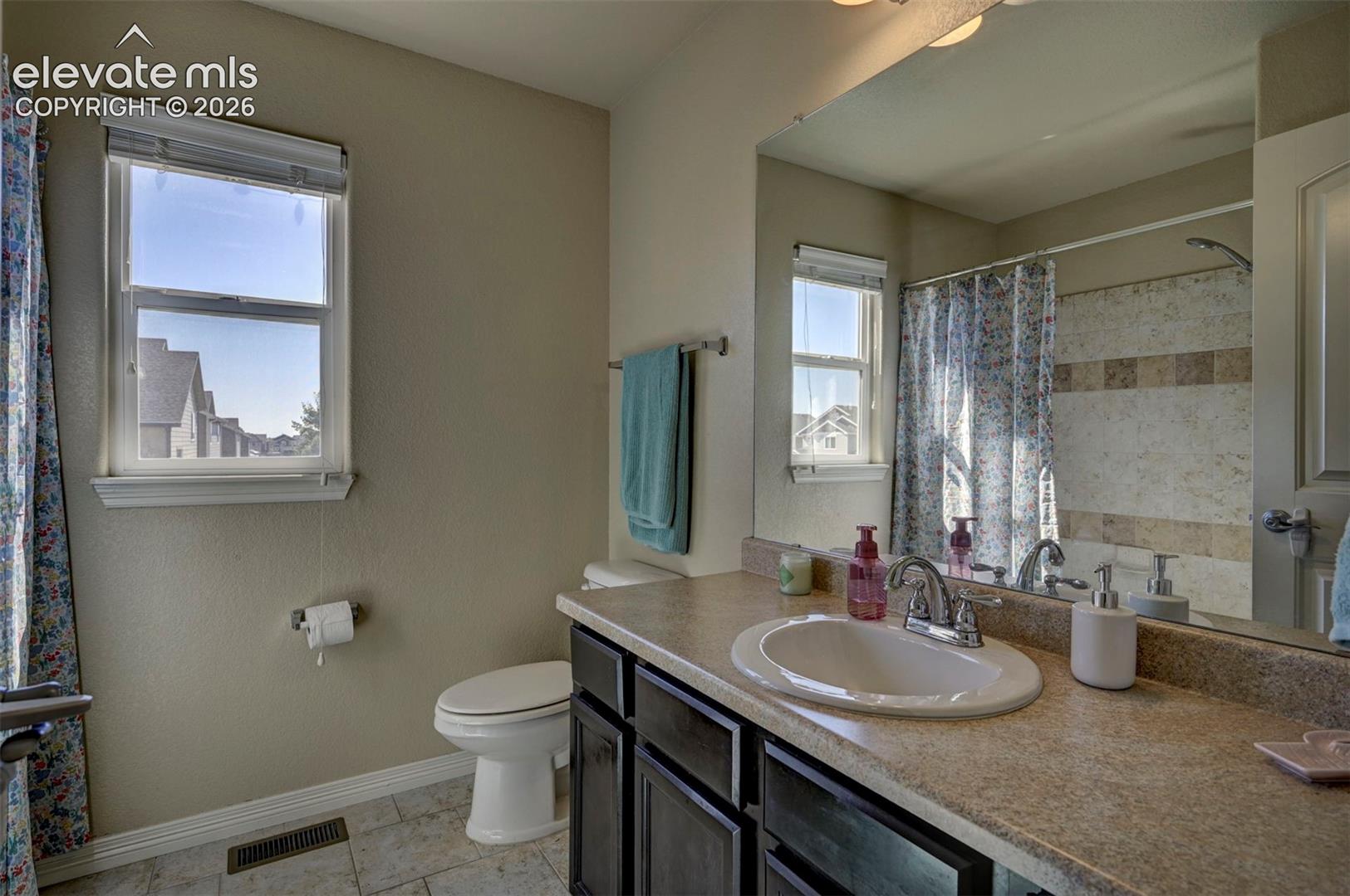 Window with blinds, vanity with integrated sink, chrome faucet, wall-mounted mirror, and tile flooring