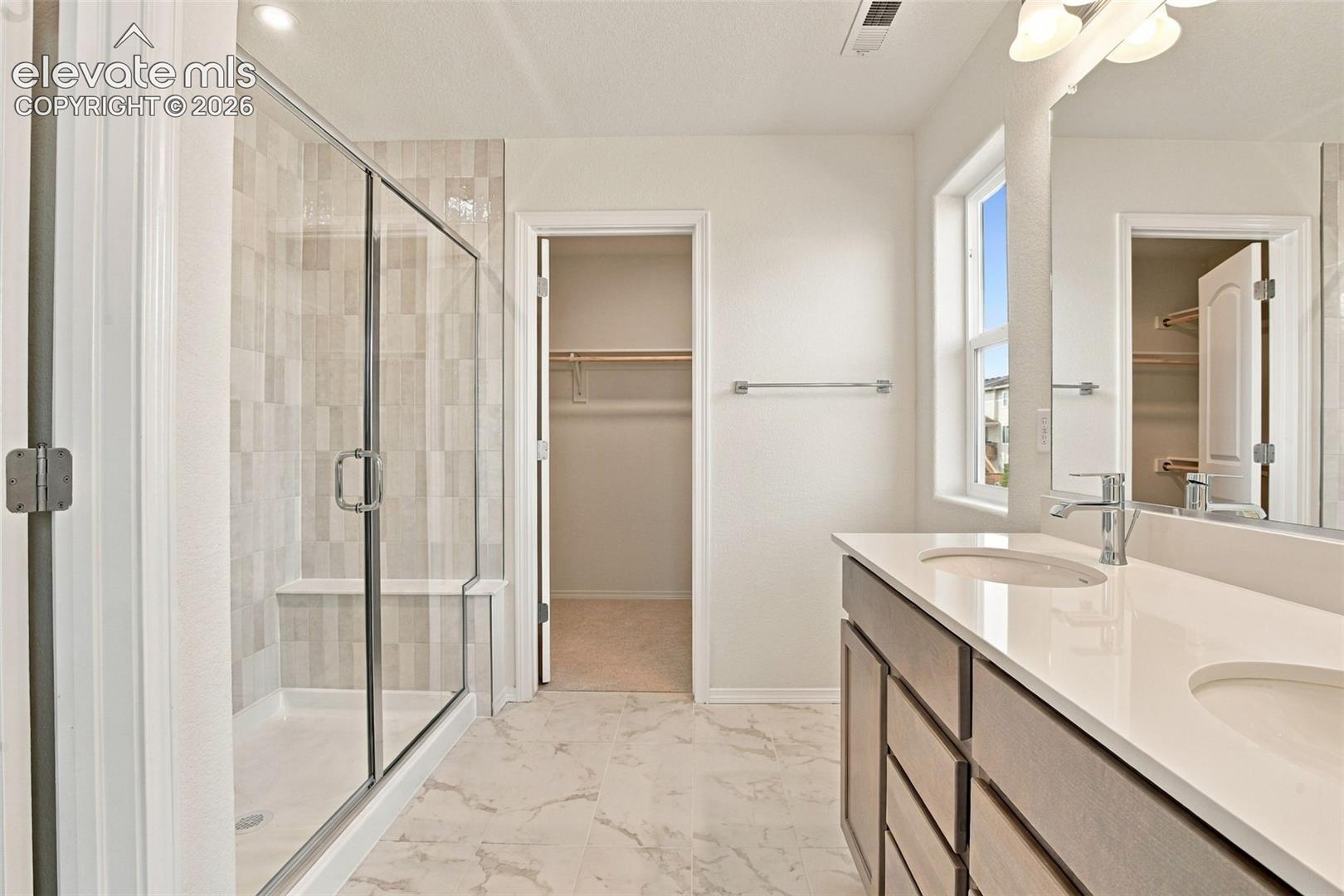 Bathroom featuring a dual-sink vanity with a solid-surface countertop, a walk-in shower with vertical tile surround and glass enclosure, large-format tile flooring, and a walk-in closet with built-in shelving