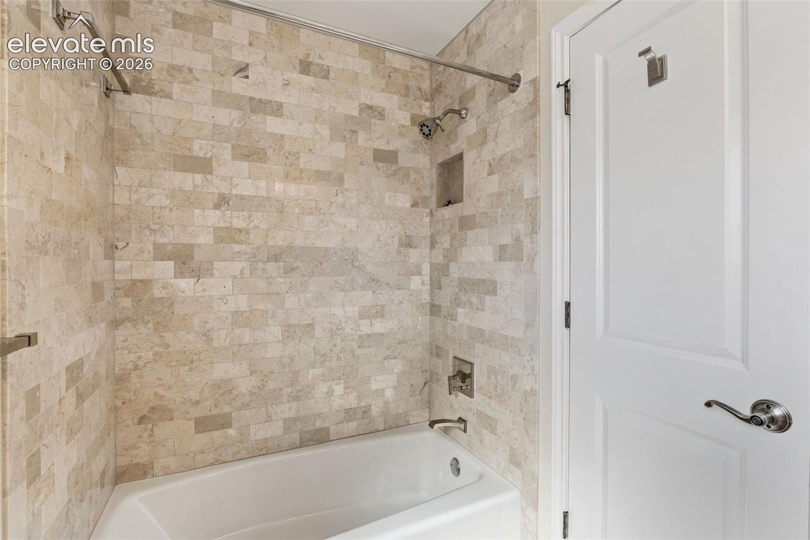 Bathtub featuring a textured stone tile surround, integrated soap niche, metallic shower head, and brushed metal faucet hardware