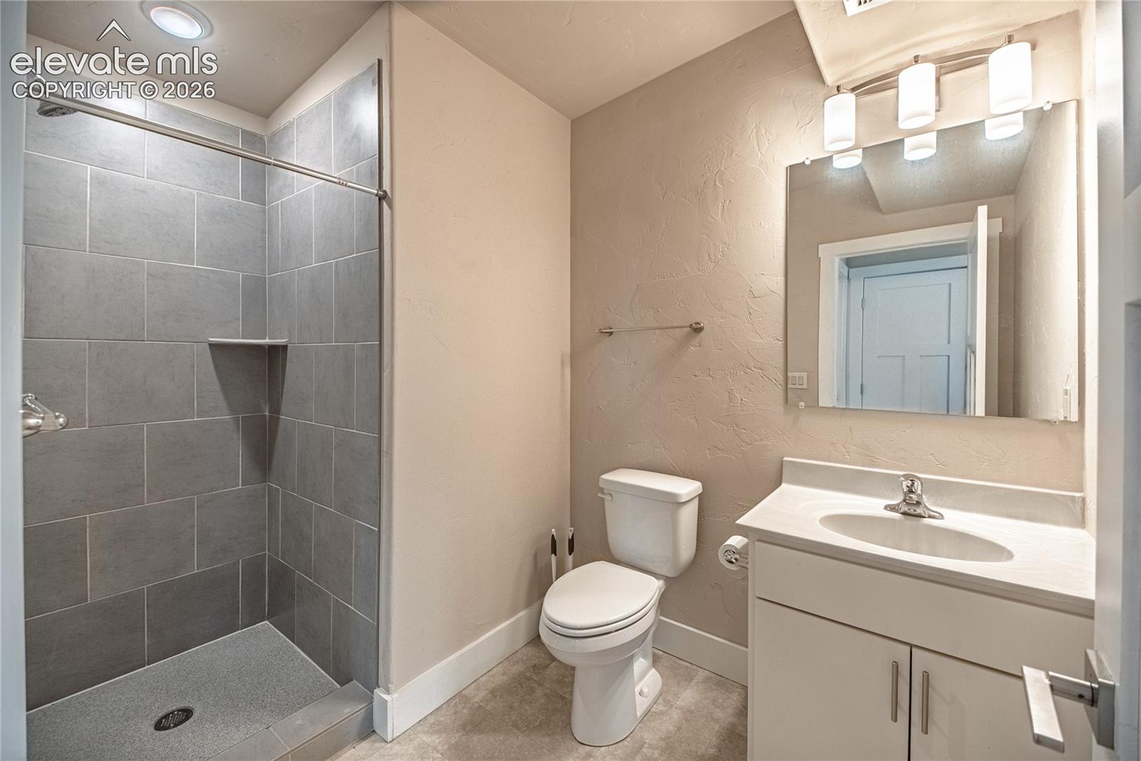 Bathroom featuring a walk-in shower with gray tile surround and integrated shelf, a vanity with a white countertop and undermount sink, a chrome faucet, and a wall-mounted mirror with overhead lighting