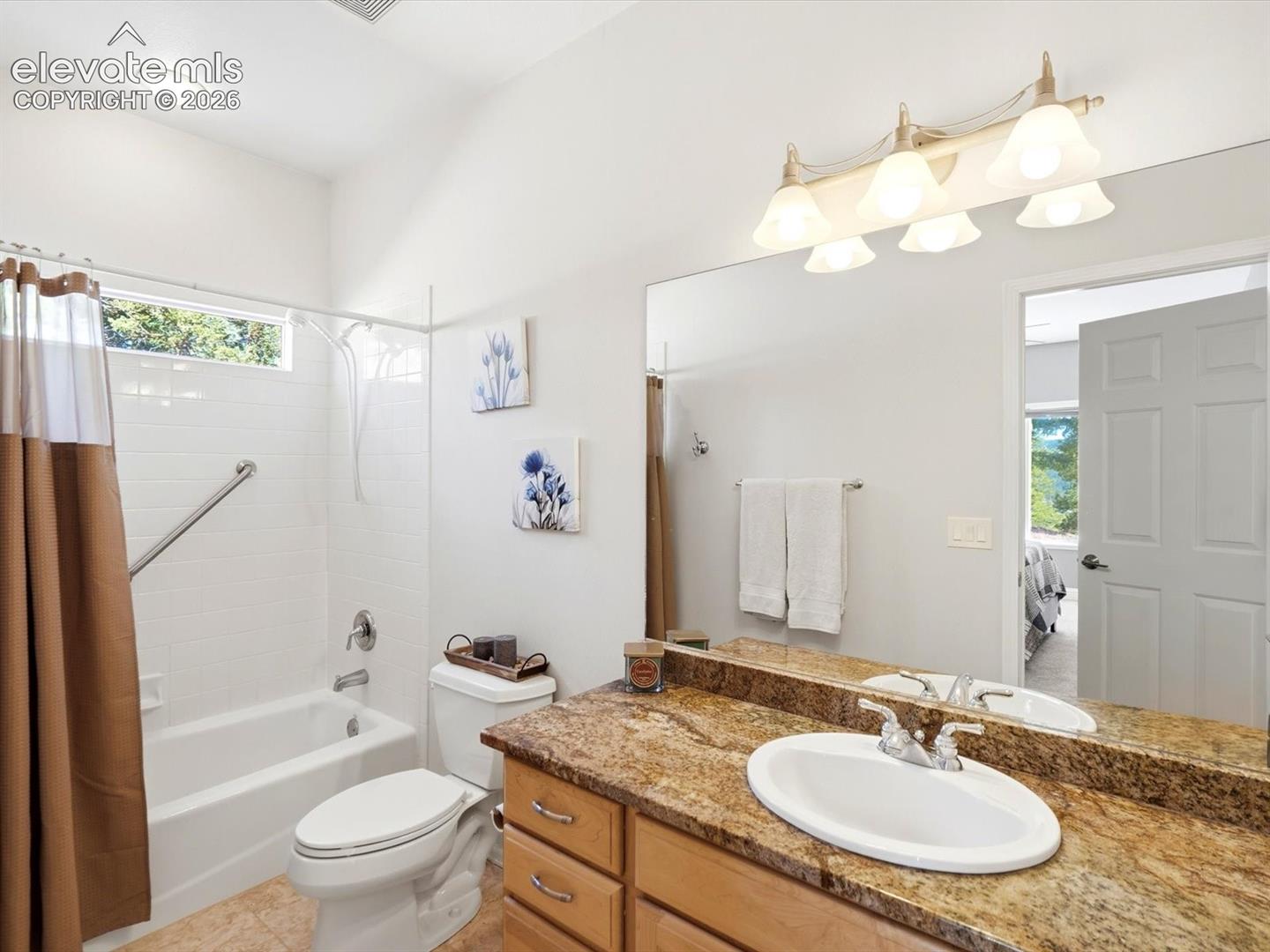 Main floor secondary en suite bathroom featuring a vanity with a stone-surface countertop, an oval under-mount sink, wooden cabinetry, and a full-width wall mirror with integrated multi-bulb lighting